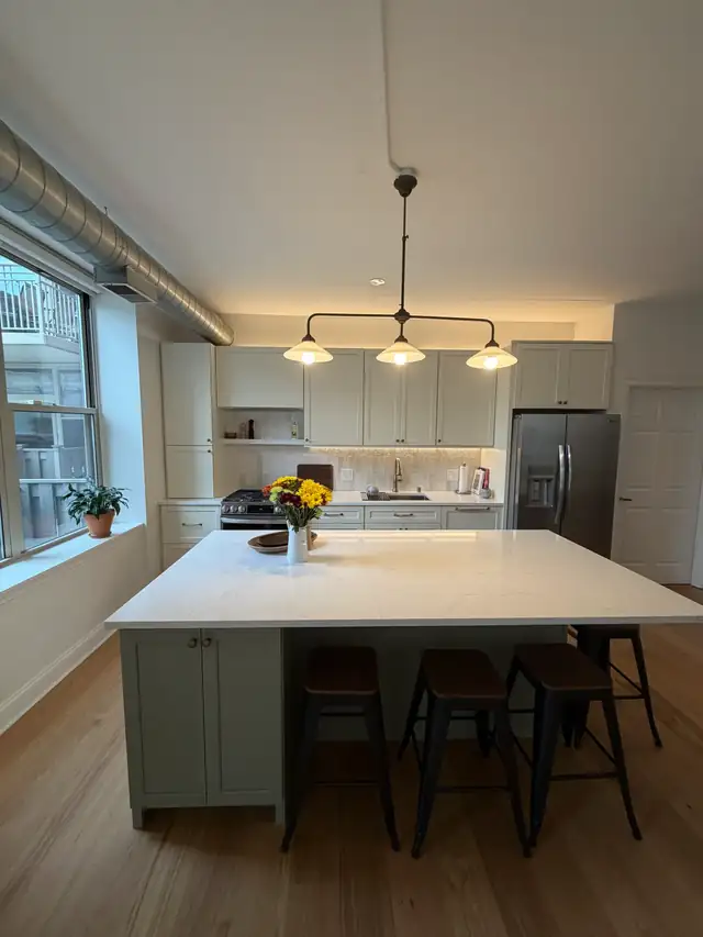 West Loop condo kitchen with a large white quartz island, sage base cabinets, black stools and a linear pendant by tall windows